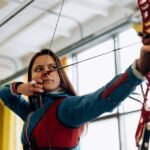 Young female archer in blue aiming a bow and arrow indoors, showcasing concentration and athleticism.