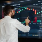 A man analyzes cryptocurrency graphs on a touchscreen monitor in a modern office setting.